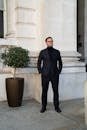 Confident man in a dark suit standing by a potted plant against a classic stone building facade.